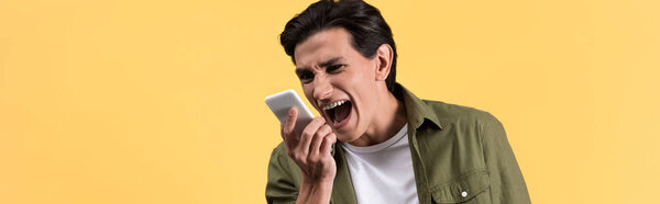 panoramic shot of aggressive young man yelling at smartphone, isolated on yellow