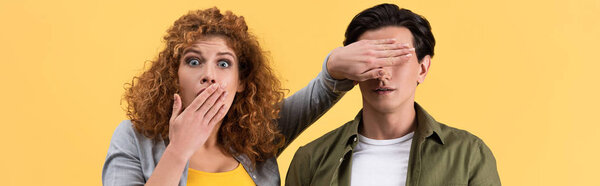 panoramic shot of shocked scared girl closing mouth while closing eyes to boyfriend, isolated on yellow