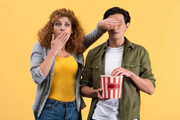scared girl watching movie while closing eyes to boyfriend with bucket of popcorn, isolated on yellow