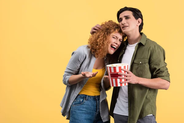 upset boyfriend hugging crying girlfriend while watching drama movie with bucket of popcorn, isolated on yellow