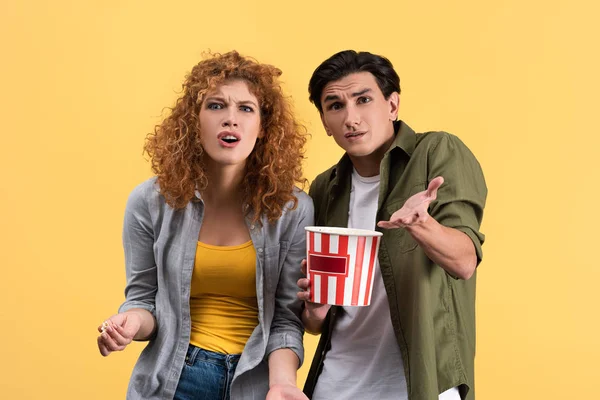 irritated couple watching bad movie with bucket of popcorn, isolated on yellow