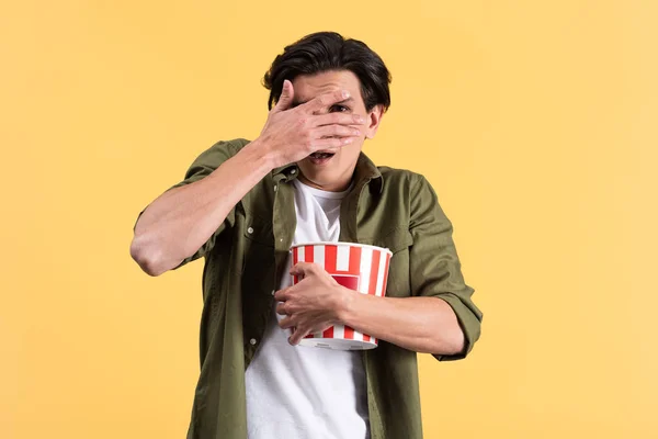 frightened young man watching horror movie, closing eyes and holding bucket of popcorn, isolated on yellow