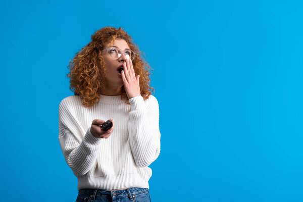 bored girl yawning, holding remote control and watching tv, isolated on blue
