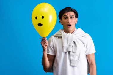 shocked young man holding yellow emotional balloon, isolated on blue