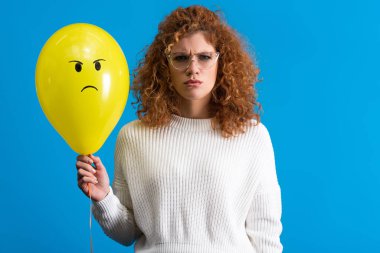 aggressive woman holding yellow balloon with angry face, isolated on blue