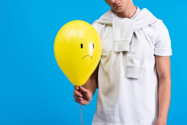 cropped view of sad man holding yellow upset balloon, isolated on blue
