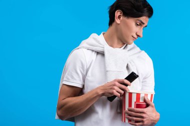 skeptical young man watching tv with remote controller and popcorn, isolated on blue