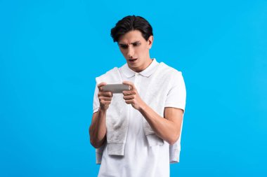 shocked young man using smartphone, isolated on blue