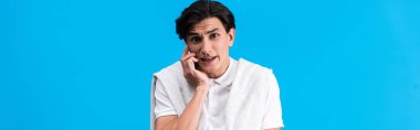 panoramic shot of irritated young man talking on smartphone, isolated on blue