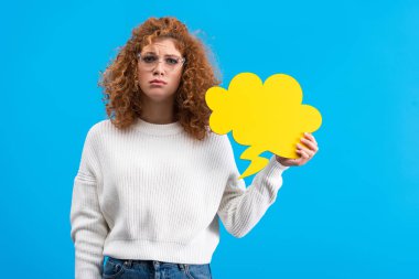 upset woman in eyeglasses holding speech bubble in shape of cloud, isolated on blue 