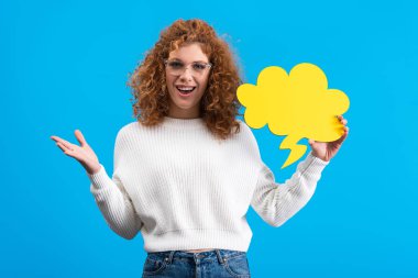 excited woman in eyeglasses holding empty speech bubble in shape of cloud, isolated on blue 