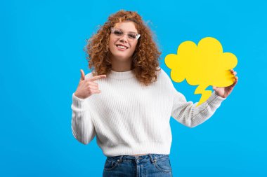 happy woman pointing at empty speech bubble in shape of cloud, isolated on blue 