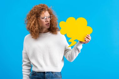 skeptical woman in eyeglasses looking at empty speech bubble in shape of cloud, isolated on blue 