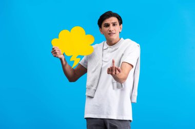 aggressive man showing middle finger while holding yellow cloud speech bubble, isolated on blue