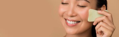 smiling beautiful asian girl with closed eyes using facial gua sha jade board isolated on beige, panoramic shot clipart