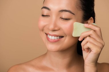 smiling beautiful naked asian girl with closed eyes using facial gua sha jade board isolated on beige clipart