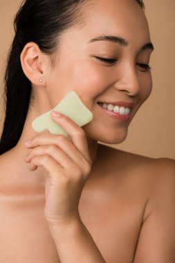 Gülümseyen güzel, çıplak Asyalı, gözleri kapalı bir kız. Yüzünde gua sha Jade tahtası var.
