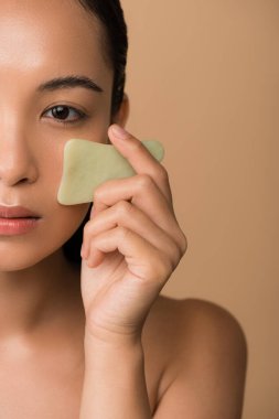Güzel, çıplak Asyalı bir kızın bej üzerine izole edilmiş yüz gua sha yeşim tahtası kullanması.