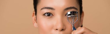 cropped view of beautiful asian girl using eyelash curlers isolated on beige, panoramic shot clipart