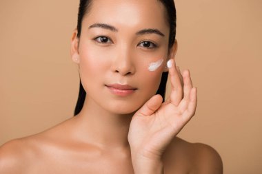 Güzel, çıplak Asyalı kız bej üzerine izole edilmiş yüz kremi sürüyor.