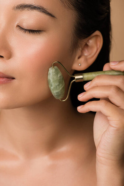close up view of beautiful naked asian girl massaging face with jade roller isolated on beige