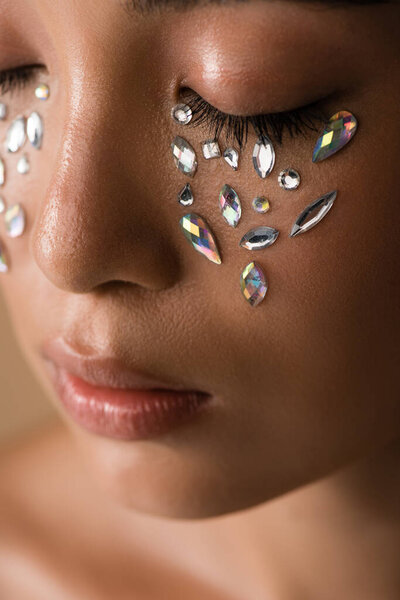 close up view of beautiful naked asian girl with closed eyes and rhinestones on face isolated on beige