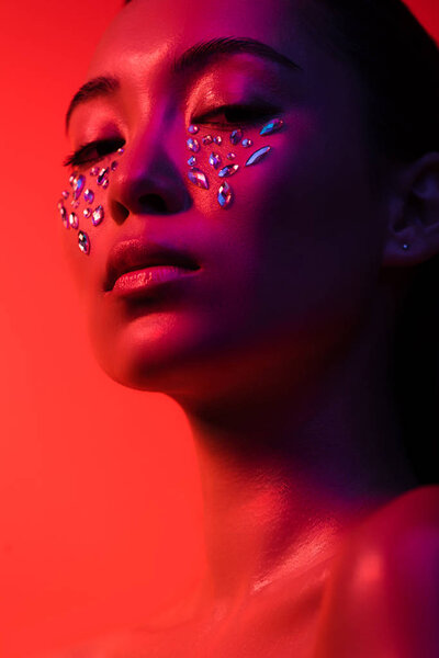 low angle view of beautiful naked asian girl with rhinestones on face isolated on red