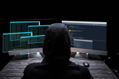 Siyah ekranda verileri olan bilgisayar monitörlerinin yanında oturan kapüşonlu hacker 'ın arka planı 