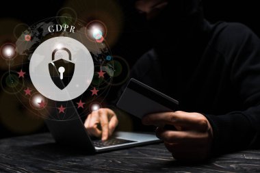 Siyah üzerine GDPR harfleri olan asma kilit yanında kredi kartı tutarken dizüstü bilgisayar kullanan maskeli hackerın seçici odağı 
