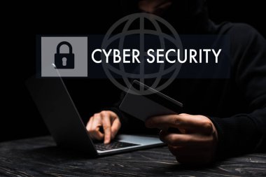 Siyah üzerine siber güvenlik harfleri yazarak kredi kartı tutarken dizüstü bilgisayar kullanan maskeli hackerın seçici odak noktası 