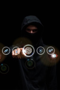 selective focus of hacker in hood pointing with finger at fingerprint web icon on black  clipart