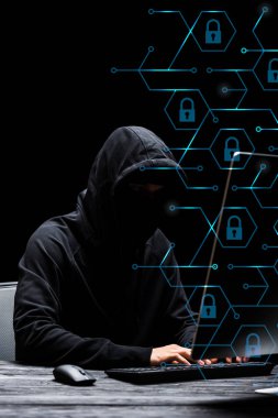 hacker in mask sitting near computer monitor and typing on computer keyboard near padlocks on black  clipart