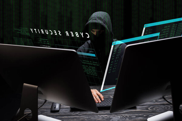 hacker in mask sitting near computer monitors and cyber security lettering on black 