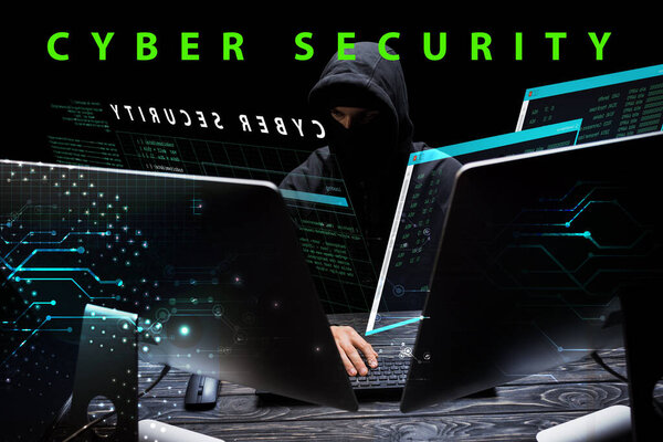 hacker in hood sitting near computer monitors near cyber security lettering on black 