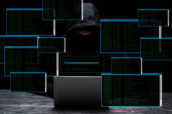 hacker in hood using laptop near screens with data on black 