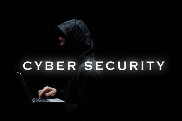 anonymous hacker in hood and mask using laptop near cyber security lettering on black 