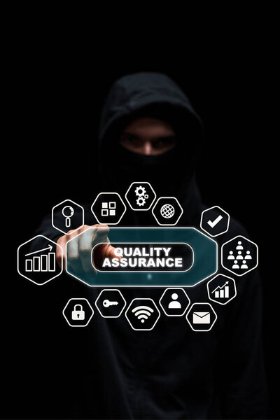 selective focus of hacker in hood pointing with finger at quality assurance lettering on black 