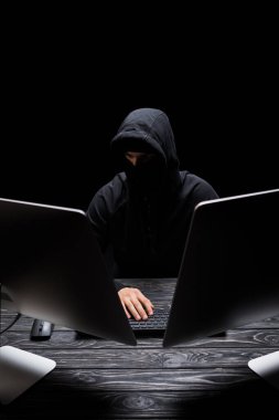 Maskeli adam bilgisayar monitörlerinin yanında siyahta izole bir şekilde oturuyor. 