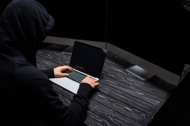 Siyah ekranda boş ekranı olan bilgisayar monitörleri yanında laptop kullanan kapüşonlu hacker 