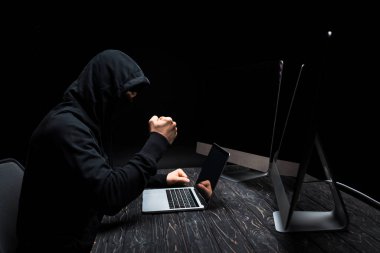 Yumruğunu dizüstü bilgisayarın yanına sıkıştırmış kızgın bir bilgisayar korsanı ve boş ekranlı bilgisayar monitörleri siyah ekranda izole edilmiş. 