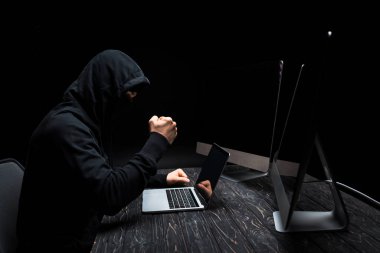 angry hacker with clenched fist near laptop and computer monitors with blank screen isolated on black  clipart