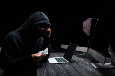 Üzgün bilgisayar korsanı dizüstü bilgisayarın yanında doku tutuyor ve boş ekranlı bilgisayar monitörleri siyah ekranda izole edilmiş. 