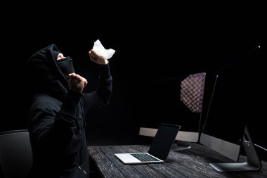 emotional hacker holding tissue near laptop and computer monitors with blank screen isolated on black  clipart