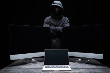Bilgisayar monitörlerinin yanında çapraz kolları olan maskeli bilgisayar korsanı ve siyah ekranlı boş ekran olan laptop. 