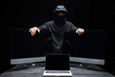 Maskeli hacker laptopun yanındaki bilgisayar monitörlerine dokunuyor ve siyah ekranda boş ekran var. 