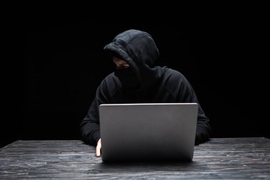 Siyah dizüstü bilgisayar kullanırken maskeli genç hacker gözünü kaçırıyor. 