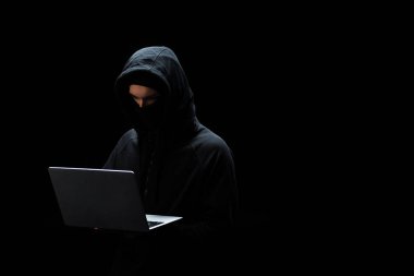Siyah üzerine izole edilmiş dizüstü bilgisayar kullanan maskeli ve başlıklı bilgisayar korsanı 