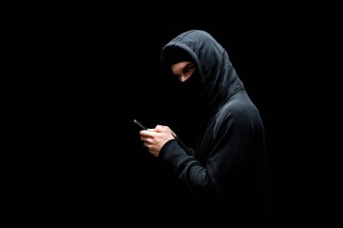 Kapüşonlu ve maskeli bir bilgisayar korsanı akıllı telefon tutuyor ve siyah kameraya bakıyor. 