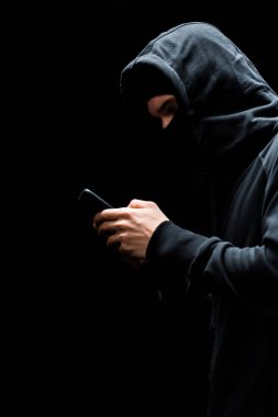 Siyah üzerine izole akıllı telefon kullanarak kapüşonlu ve maskeli bilinmeyen bir hacker. 