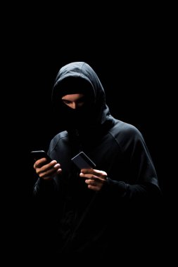 Kapüşonlu ve maskeli genç bir bilgisayar korsanı akıllı telefon kullanıyor ve elinde siyah kart tutuyor. 